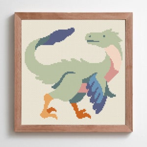 May include: A framed cross-stitch artwork depicting a stylized dinosaur in green, blue, pink, and orange. The dinosaur is set against a cream background and framed in a light brown wooden frame. The piece has a handmade aesthetic, with visible stitches.