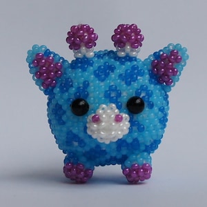 以下が含まれることがあります： 白のアクセントが付いた青と紫のビーズでできたキリンの玩具。キリンは丸い体と小さな脚をしています。