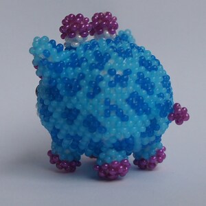 以下が含まれることがあります： 青と紫のビーズでできた小さな動物の置物。置物は丸い体と4本の足、そして頭の上に2つの小さな紫色のビーズの耳がついています。