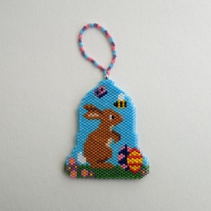 Könnte beinhalten: Ein blauer Perlen-Anhänger in Form einer Glocke mit einem braunen Kaninchen, einer Biene und einem Osterei darin. Der Anhänger hat eine rosa und violette Schlaufe zum Aufhängen.