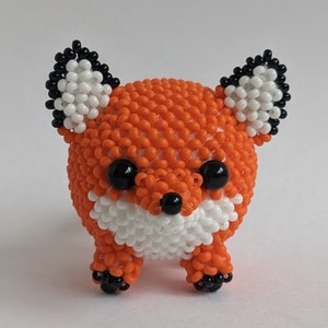 Könnte beinhalten: Eine kleine orangefarbene und weiße Perlenfigur eines Fuchses mit schwarzen Augen und einer schwarzen Nase.