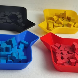 Puede incluir: Cuatro bandejas pequeñas de forma cuadrada, cada una de un color diferente: negro, amarillo, azul y rojo. Cada bandeja contiene una pila de pequeños bloques de plástico de colores.