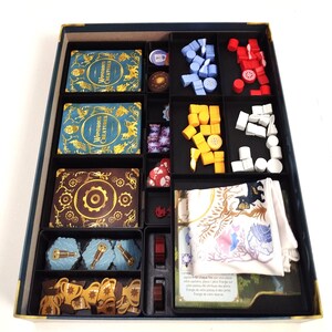 Op de afbeelding: Een bordspel organizer met diverse componenten. De doos bevat kaarten met gouden en turquoise ontwerpen, houten tokens in blauw, rood, geel en wit, en een witte doek met een bloemenpatroon. Het spel heet "Wondrous Creatures".