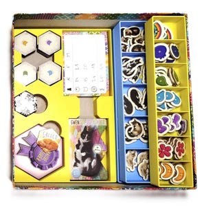 Può includere: Una scatola di gioco da tavolo con scomparti gialli, blu e fantasia. La scatola contiene pedine, carte e gettoni a tema gatto. I pezzi del gioco includono illustrazioni di gatti, forme di fiori e forme di mezzaluna. La scatola presenta anche il testo "CALICO MAITRE TURIER GWEN".