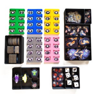 Puede incluir: Un conjunto de cajas de almacenamiento de plástico de colores con compartimentos para organizar pequeñas piezas de juego. Las cajas están llenas de varios componentes de juego, incluidos dados, fichas y cartas.
