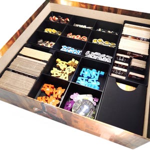 Peut inclure: Une boîte en bois avec un design brun et or, contenant une variété de pièces de jeu et de cartes. La boîte est divisée en compartiments pour un rangement organisé. Les pièces de jeu sont en plastique et en bois, et les cartes sont en carton.