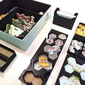 Può includere: Un set organizzatore per giochi da tavolo con vassoi e contenitori in plastica nera. Il set include una scatola grande con divisori, un porta carte e diversi vassoi più piccoli contenenti tessere esagonali, monete d'oro e gettoni con pesci.