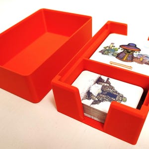 Puede incluir: Caja de almacenamiento de plástico naranja para juegos de cartas con dos compartimentos para cartas. La caja tiene una tapa y un divisor para separar las cartas.