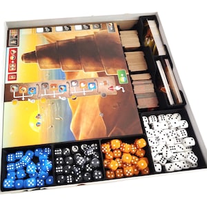 May include: A board game organiser with a game board, cards, and dice. The game board depicts a desert landscape with a large structure. Dice are blue, black, orange, and white. The organiser is divided into compartments for game components.
