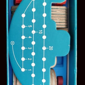 May include: A teal wooden puzzle game featuring numbered holes and a curved path. The game includes small wooden discs and red cylindrical pieces, all contained within a blue box.