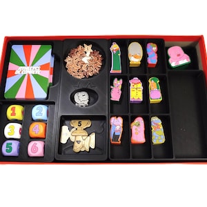 Puede incluir: Un juego de mesa en una caja roja con un inserto de plástico negro que contiene una baraja de cartas con el texto "Athlètes de Compète" y un patrón de rayos, seis dados numerados, fichas de estrellas de madera, trofeos dorados y figuras coloridas.