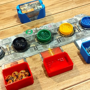 Può includere: Un organizer per giochi da tavolo con vari componenti. Include un porta carte blu, diversi contenitori colorati e supporti circolari in verde, giallo, nero e blu. Il tabellone è grigio con un design futuristico e i componenti sono disposti su una superficie di legno.