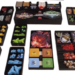 Puede incluir: Bandejas organizadoras negras llenas de componentes de juego, incluyendo dados, fichas y cartas. Las bandejas están dispuestas para contener varias piezas de juego, con diferentes colores y formas. Los componentes son para un juego de mesa.