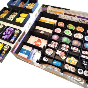 Könnte beinhalten: Ein schwarz-weißer Spielorganizer mit mehreren Fächern zum Verstauen von Spielsteinen. Der Organizer hat eine Vielzahl von Fächern zum Verstauen von Spielsteinen unterschiedlicher Größe, darunter kleine Token, Würfel und Karten. Der Organizer ist aus robustem Kunststoff gefertigt und so konzipiert, dass er in eine Standard-Spielbox passt.