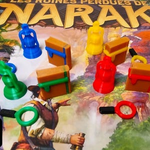 May include: A board game with the title "LES RUINES PERDUES DE NARAK" in gold lettering. The game pieces are colorful, including blue, green, yellow, and red figures, and brown rectangular pieces with colored bands. A magnifying glass is also visible.
