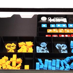 Op de afbeelding: Een zwarte speldoos met compartimenten met gele, blauwe, oranje en rode spelstukken. De doos bevat de tekst "AMBITION" op een witte kaart. De spelstukken hebben verschillende vormen.