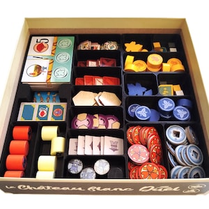 May include: A beige game box with black compartments filled with game pieces. The pieces include cards, wooden tokens, and colorful circular and rectangular pieces. The box has the text "le Château blanc Duel" on the front.