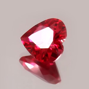 May include: A heart-shaped, faceted, red gemstone. The gem is highly reflective, with multiple facets catching the light. The gemstone is set against a light background, with its reflection visible below.