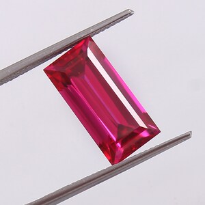 May include: A vibrant, deep red rectangular gemstone with a faceted cut. The gemstone is held by a pair of tweezers.