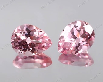 May include: Two pear-shaped pink gemstones with facets. The gemstones are on a white surface.