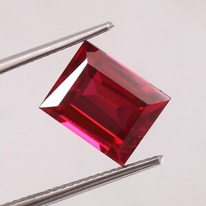 May include: A deep red emerald cut gemstone held by tweezers. The gemstone is a vibrant red color and has a rectangular shape.