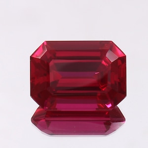May include: A faceted, rectangular ruby gemstone with a deep red hue. The gem is cut in an emerald shape, showcasing its facets and internal reflections. The stone is displayed on a reflective surface, enhancing its brilliance.
