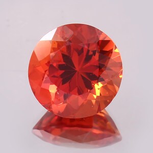May include: A round, faceted gemstone with a vibrant orange-red hue. The gem is highly reflective, with a complex internal pattern visible. The stone is set against a light background, with its reflection visible below.