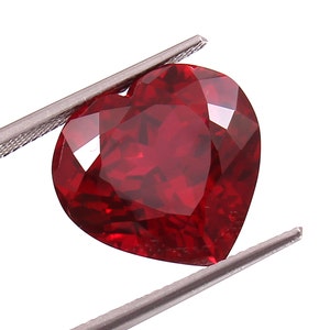 Può includere: Una pietra preziosa di colore rosso scuro a forma di cuore con taglio a faccette. La pietra preziosa è tenuta da una pinzetta.