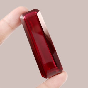 May include: A deep red gemstone with a rectangular cut and a high polish. The gemstone is held between two fingers.