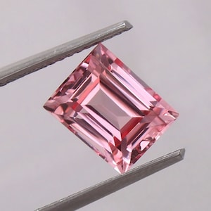 May include: A pink gemstone with a rectangular emerald cut. The gemstone is held by a pair of tweezers.