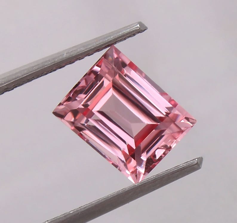May include: A pink gemstone with a rectangular emerald cut. The gemstone is held by a pair of tweezers.