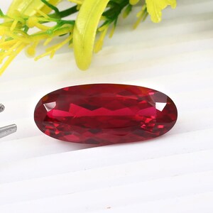 May include: A deep red oval-shaped gemstone with a faceted cut. The gemstone is set against a white background.