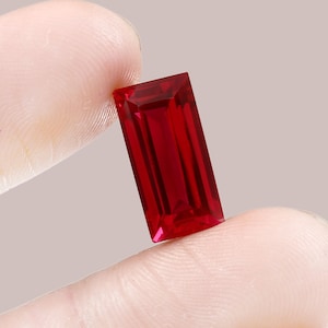 May include: A deep red rectangular gemstone with a faceted cut. The gemstone is held between two fingers.