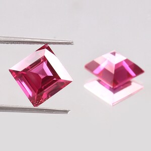 Könnte beinhalten: Zwei quadratisch geschliffene, facettierte Edelsteine in leuchtendem Pink. Ein Stein wird von einer Pinzette gehalten, während der andere auf einer reflektierenden Oberfläche liegt und seine geometrische Form und Farbe zeigt.