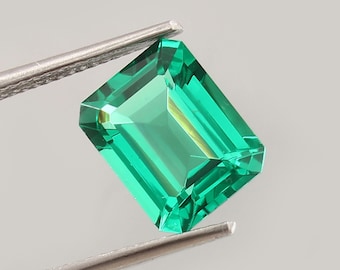 Könnte beinhalten: Ein leuchtend grüner, smaragdgeschliffener Edelstein, gehalten von einer silbernen Pinzette. Der rechteckige Stein hat Facetten, die das Licht reflektieren. Der Hintergrund ist ein neutrales Grau.