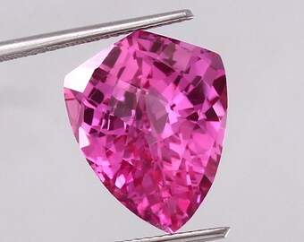 May include: A vibrant pink gemstone with a faceted, shield-shaped cut. The gemstone is held by a pair of tweezers.