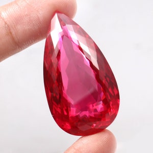 May include: A pear-shaped, faceted gemstone with a deep pink hue. The gemstone is held in a hand, showcasing its brilliance and clarity.