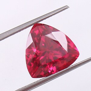 May include: A deep red triangular gemstone with a faceted cut, held by a pair of tweezers.