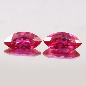 May include: Two bright pink marquise-shaped gemstones, likely rubies, are displayed on a white surface. The gemstones are faceted and reflect light.