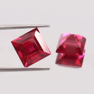 May include: Two vibrant, deep red square-cut gemstones with faceted surfaces. One gemstone is held by silver-toned metal tweezers, while the other rests nearby, displaying a clear reflection of its geometric shape and brilliant crimson colour.