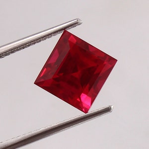 Könnte beinhalten: Ein leuchtend roter quadratischer Edelstein, der von einer Pinzette gehalten wird. Der Edelstein ist facettiert und hochglanzpoliert.