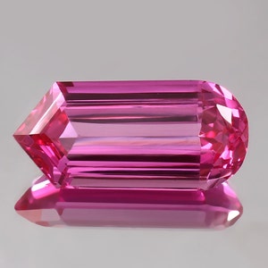 Puede incluir: Una gema rosa vibrante con forma rectangular facetada. La gema tiene una superficie lisa y pulida y está tallada en un diseño único y alargado.