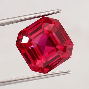May include: A vibrant red gemstone with a square emerald cut. The gemstone is held by a pair of tweezers.