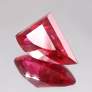 May include: A faceted, vibrant red gemstone with a unique, angular cut. The gem's surface reflects light, creating a brilliant display of color and sparkle. The gem is displayed on a reflective surface.