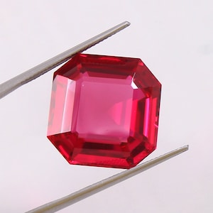Könnte beinhalten: Ein leuchtend roter Edelstein mit quadratischem Smaragdschliff. Der Edelstein wird von einer Pinzette gehalten.