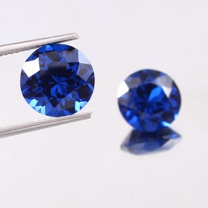 Op de afbeelding: Twee ronde, gefacetteerde, blauwe edelstenen. De edelstenen worden in een pincet gehouden en bevinden zich op een wit oppervlak.