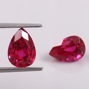 Peut inclure: Deux pierres précieuses rubis en forme de poire, l'une tenue par une pince, d'une couleur rouge foncé et d'un éclat brillant.