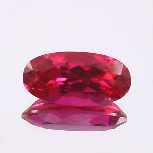 Puede incluir: Una gema ovalada de color rojo vibrante con un corte facetado. La gema refleja la luz, creando un efecto brillante.