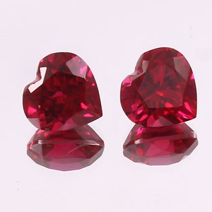 May include: Two heart-shaped ruby gemstones, each with a brilliant red color and a faceted cut. The gemstones are displayed on a white surface.