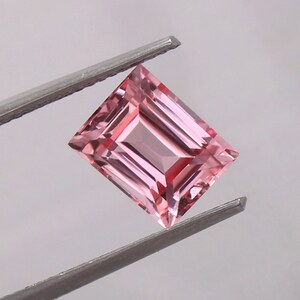 May include: A pink gemstone with a rectangular emerald cut. The gemstone is held by a pair of tweezers.